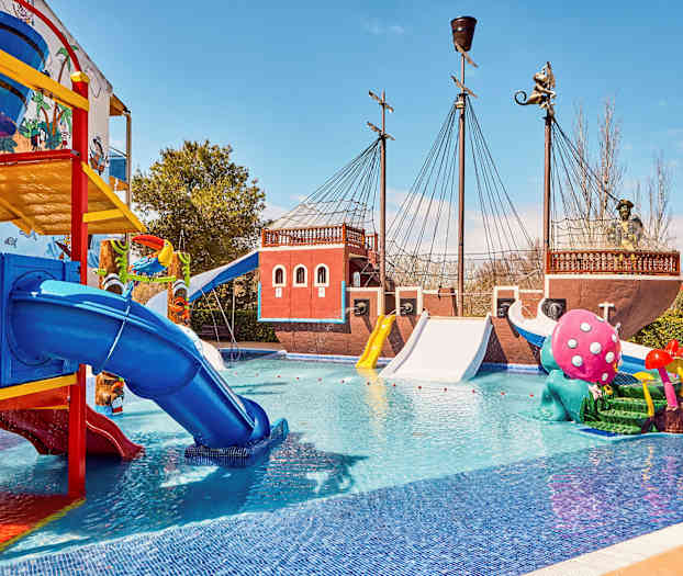 Kuva vesipuistosta, jossa on merirosvoteemainen laiva, värikkäitä liukumäkiä ja leikkiveistoksia altaassa. (Tekoälyn luoma teksti)