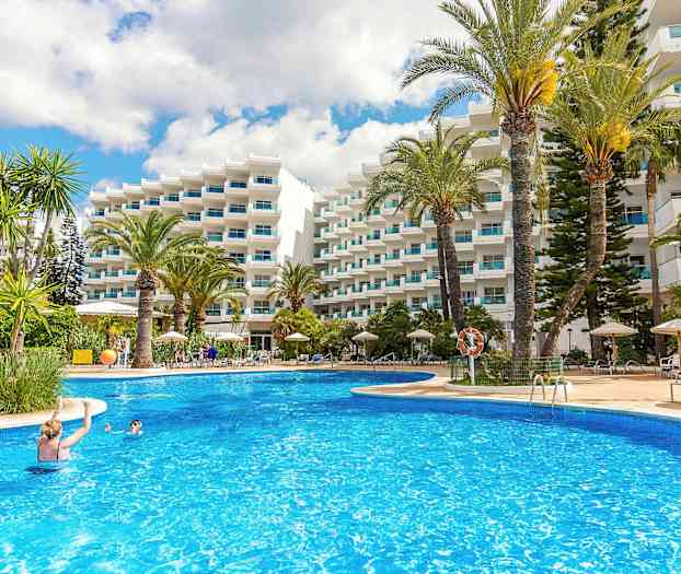 Ulkouima-allas, jota ympäröivät aurinkotuolit, aurinkovarjot ja korkeat palmut, valkoisen parvekkeellisen rakennuksen edessä. (Tekoälyn luoma teksti)