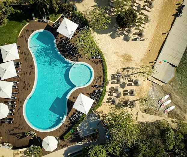 Ilmakuva uima-altaasta, jota ympäröivät aurinkotuolit ja varjot, vieressä ranta ja laituri. (Tekoälyn luoma teksti)