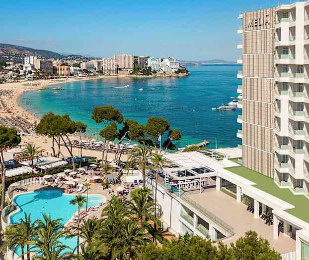 Ranta, jossa on kirkasta sinistä vettä, ympäröitynä hotellirakennuksilla, puilla ja uima-altaalla. (Tekoälyn luoma teksti)