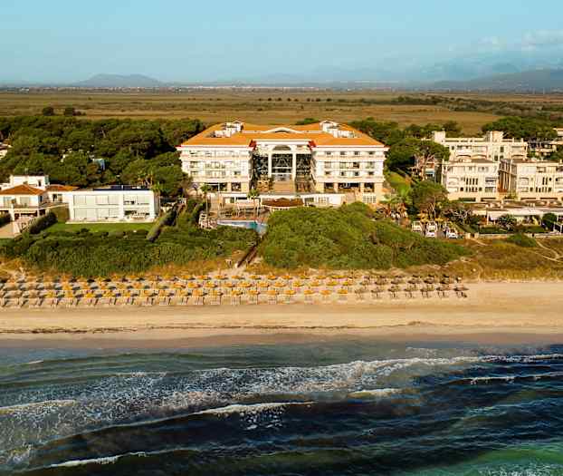 Näkymä rannalle, jossa aurinkotuoleja ja aurinkovarjoja hotellin edessä, ympärillä kasvillisuutta. (Tekoälyn luoma teksti)