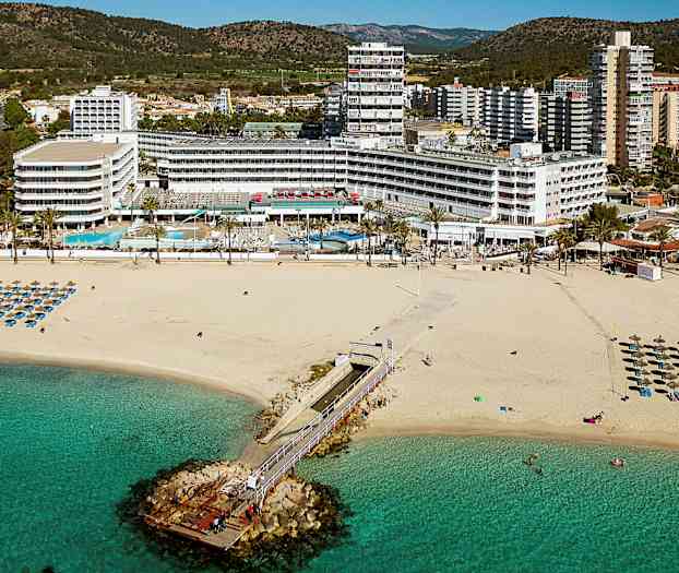 Kuva hiekkarannasta, jossa aurinkotuoleja ja aurinkovarjoja, laituri joka johtaa merelle, sekä hotellirakennuksia taustalla. (Tekoälyn luoma teksti)