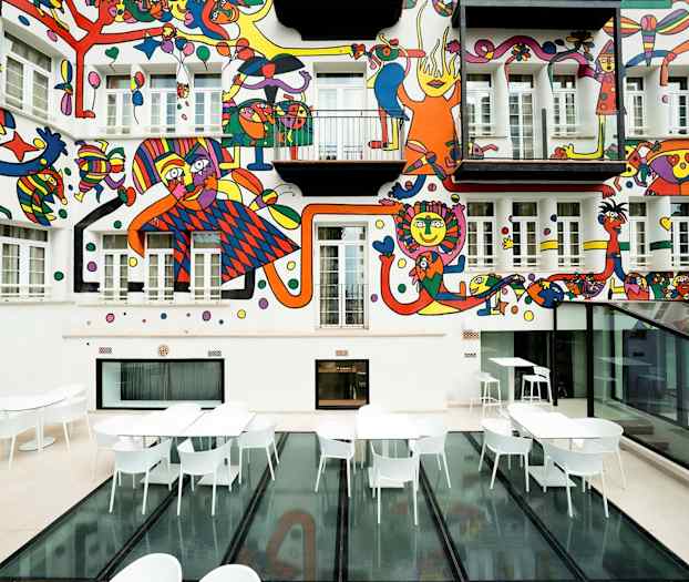 Sisäpiha, jossa on valkoisia pöytiä ja tuoleja sekä värikäs seinämaalaus talon julkisivussa. (Tekoälyn luoma teksti)