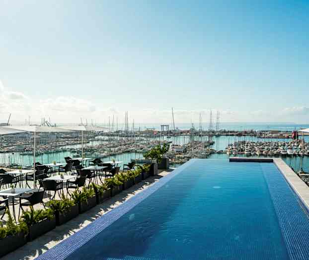Infinity-allas terassilla, jolta on näkymä satamaan, joka on täynnä veneitä, ja merelle taustalla. Altaan vieressä on istumapaikkoja aurinkovarjojen alla. (Tekoälyn luoma teksti)