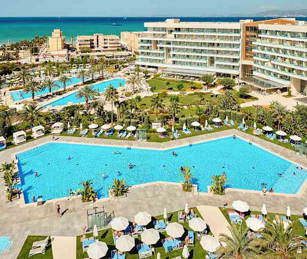 Näkymä lomakohteesta, jossa on uima-altaita, aurinkotuoleja ja ympäröivät hotellirakennukset sekä meri taustalla. (Tekoälyn luoma teksti)