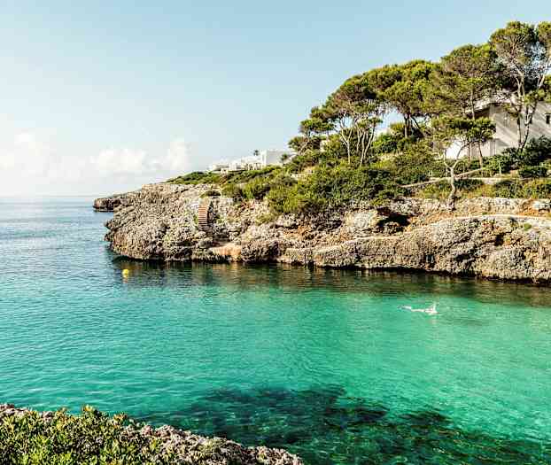 Rannikko kallioineen ja kirkas turkoosi vesi, talo näkyy puiden joukossa. (Tekoälyn luoma teksti)