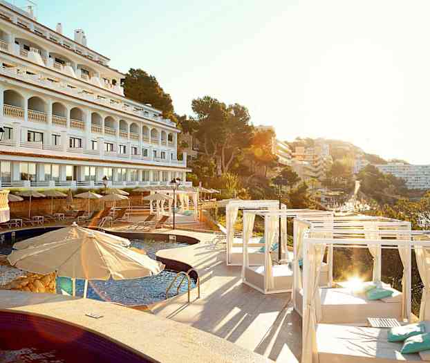 Valkoinen hotellin julkisivu, jossa kaaret, uima-allas ja aurinkotuoleja varjoineen sekä päivänvuoteita, ympärillä puita ja rakennuksia. (Tekoälyn luoma teksti)