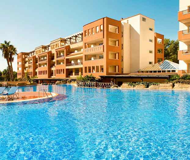 Kuvassa on rakennus suuren ulkouima-altaan vierellä, jossa on sinistä vettä ja aurinkotuoleja altaan reunalla. Taustalla näkyy muutamia palmuja. (Tekoälyn luoma teksti)