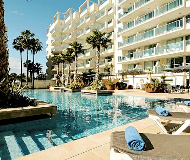 Kuva hotellin uima-altaasta, jossa on aurinkotuoleja ja palmuja modernin rakennuskompleksin ympäröimänä. (Tekoälyn luoma teksti)