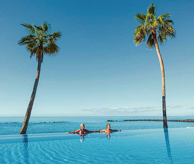 Kaksi ihmistä infinity-altaassa, josta on näkymä merelle ja kaksi palmua. (Tekoälyn luoma teksti)