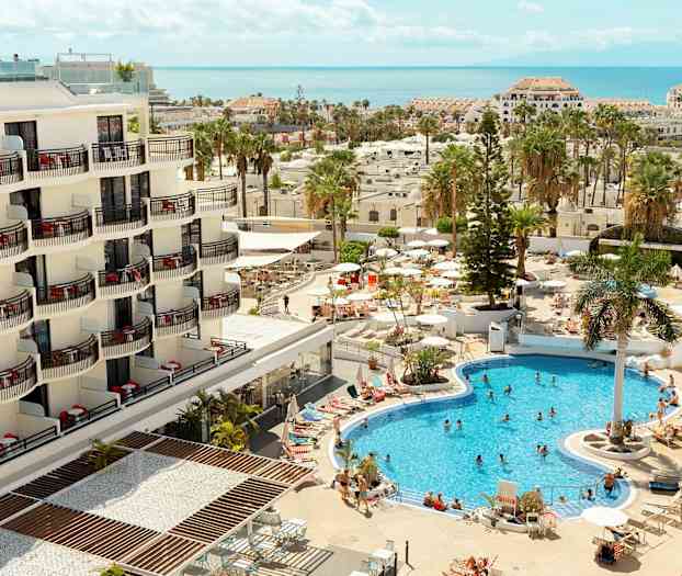 Hotelli, jossa on parvekkeet ja uima-allasalue vierailijoineen ja aurinkovarjoineen. (Tekoälyn luoma teksti)