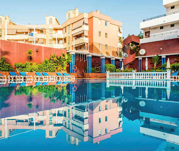 Ulkoallas, jota ympäröivät aurinkotuolit ja vihreät kasvit, taustalla rakennuksia. (Tekoälyn luoma teksti)