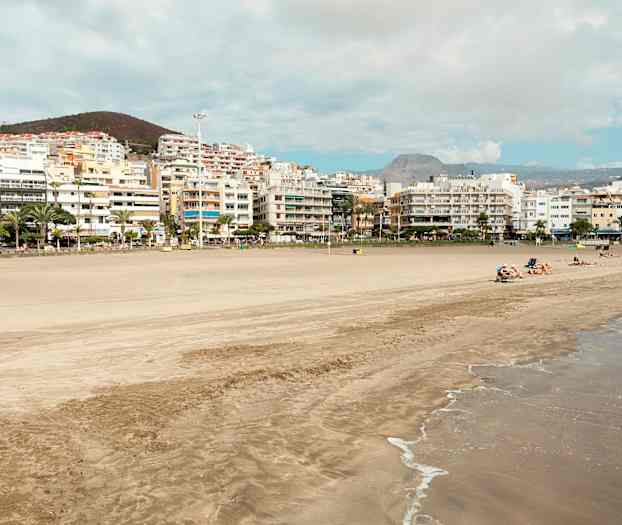 Ranta, jossa hiekkaa ja aaltoja, taustalla rakennuksia ja kukkuloita. (Tekoälyn luoma teksti)