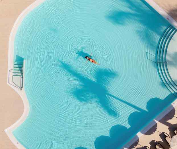 Nainen kelluu hotellin altaassa, ympärillään aurinkovarjoja ja palmuja. (Tekoälyn luoma teksti)