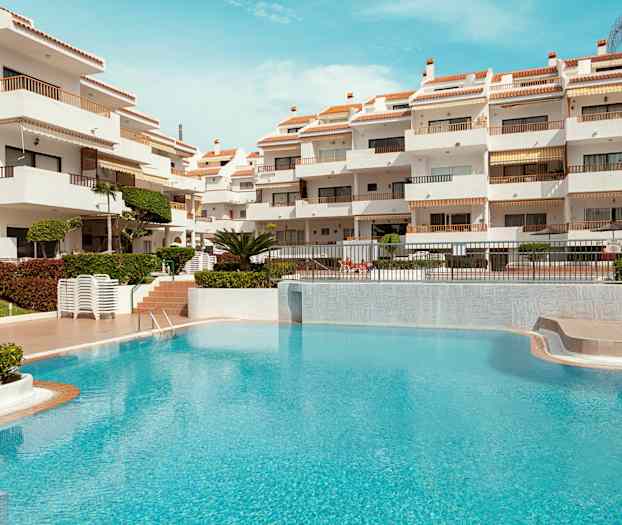 Valkoinen rakennus, jossa on terasseja, ja etualalla turkoosi uima-allas. (Tekoälyn luoma teksti)