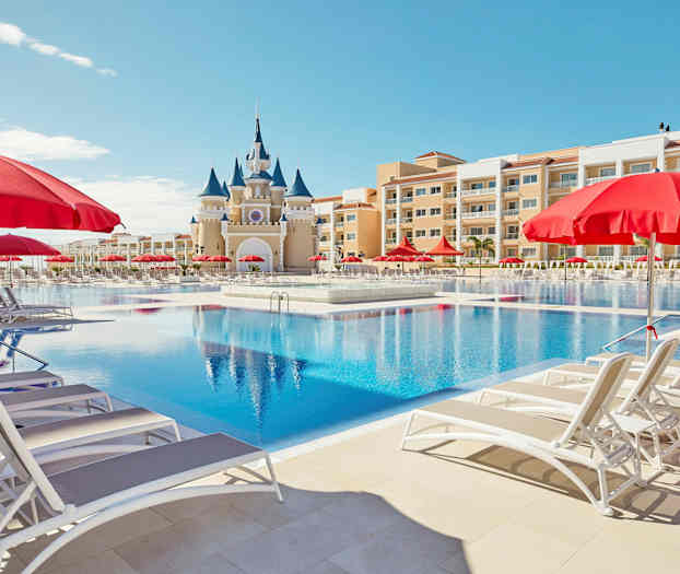 Suuri uima-allas, aurinkotuoleja ja punaisia varjoja sekä taustalla linnalta näyttävä rakennus. (Tekoälyn luoma teksti)