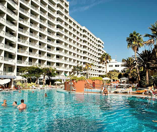 Suuri hotellirakennus, jossa on monia parvekkeita, ympäröity uima-altaalla ja palmuilla. (Tekoälyn luoma teksti)