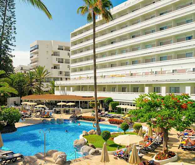 Hotelli, jonka edustalla on uima-allas, ympärillä aurinkotuoleja ja aurinkovarjoja sekä vehreyttä. (Tekoälyn luoma teksti)