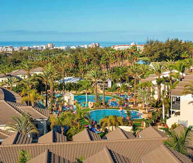 Näkymä lomahotelliin, jossa on palmuja, uima-altaita ja rakennuksia, taustalla näkyy meri ja kaupunki. (Tekoälyn luoma teksti)