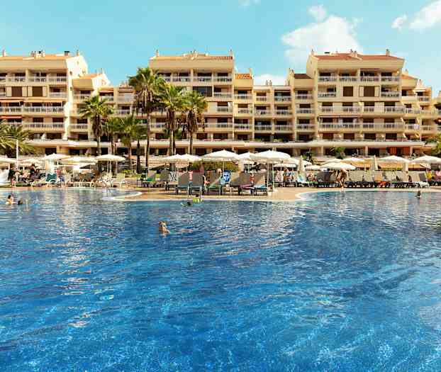 Suuri uima-allas, jota ympäröivät aurinkotuolit ja varjot, sijaitsee usean kerroksen hotellirakennuksen edessä. (Tekoälyn luoma teksti)