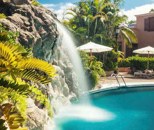 Vesiputous virtaa uima-altaaseen, ympäröitynä vehreydellä ja aurinkotuoleilla. (Tekoälyn luoma teksti)