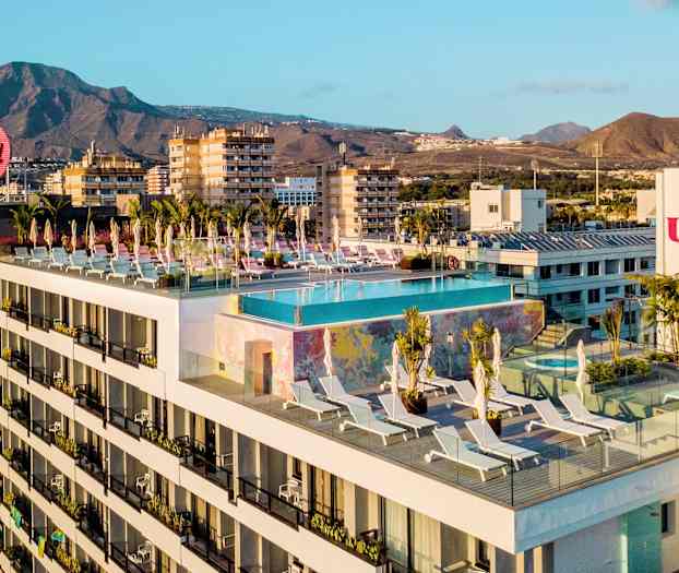 Hotellirakennus, jonka kattoterassilla on infinity-allas ja aurinkotuoleja, taustalla vuoria ja kaupunkeja. (Tekoälyn luoma teksti)