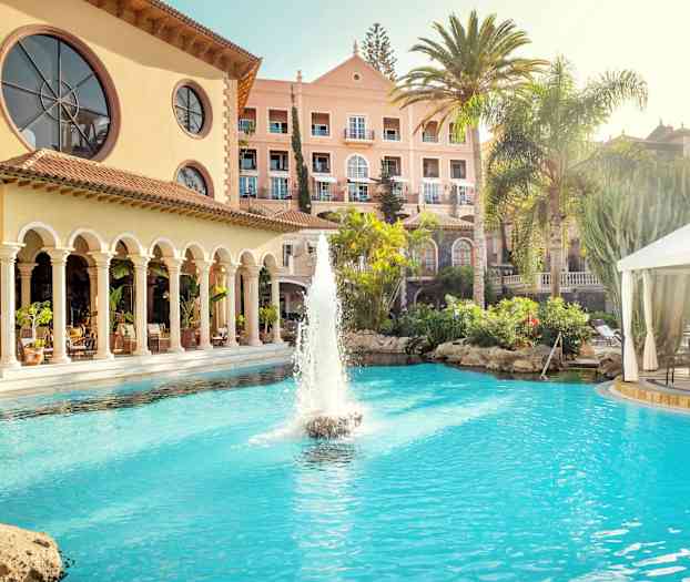 Hotellirakennus, jonka edessä on uima-allas ja suihkulähde, ympäröitynä palmuilla ja kasvillisuudella. (Tekoälyn luoma teksti)