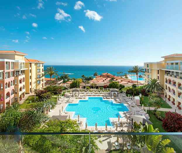 Kuva hotellialueesta, jossa on uima-allas, jota ympäröivät aurinkotuolit ja aurinkovarjot. Hotellin rakennukset sijaitsevat molemmilla puolilla, ja taustalla näkyy meri. (Tekoälyn luoma teksti)