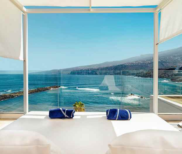 Näköala terassilta, jossa on aurinkotuoleja, lasikaide, kaksi sinistä pyyhettä ja martini-lasi etualalla, taustalla meri, kallioita ja vuoria. (Tekoälyn luoma teksti)