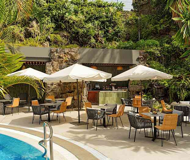 Ulkoterassi uima-altaan vieressä sisältäen pöytiä, tuoleja ja aurinkovarjoja. Taustalla vihreää kasvillisuutta ja pieni baari. (Tekoälyn luoma teksti)