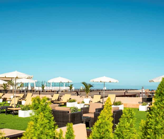 Kattoterassi, jossa on aurinkotuoleja, varjoja ja kasveja kirkkaan sinisen taivaan alla. (Tekoälyn luoma teksti)