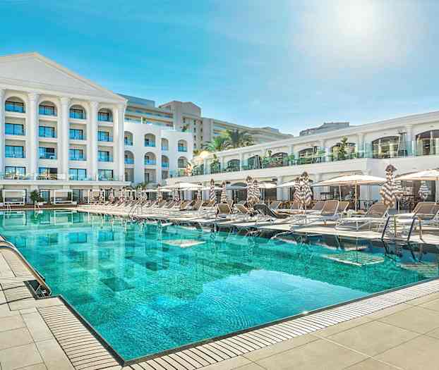 Hotellirakennus, jonka edessä on suuri uima-allas ja aurinkotuolit. (Tekoälyn luoma teksti)