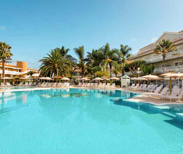 Ulkoallas, jossa aurinkotuoleja ja varjoja, ympärillä palmuja ja rakennuksia. (Tekoälyn luoma teksti)