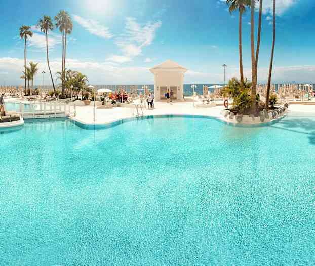 Suuri ulkoallas, jota ympäröivät aurinkotuolit ja palmut, taustalla meri. (Tekoälyn luoma teksti)