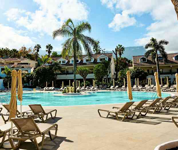 Hotellikompleksi, jossa on uima-allas, aurinkotuoleja ja -varjoja. Rakennuksia palmun ympäröimänä. (Tekoälyn luoma teksti)