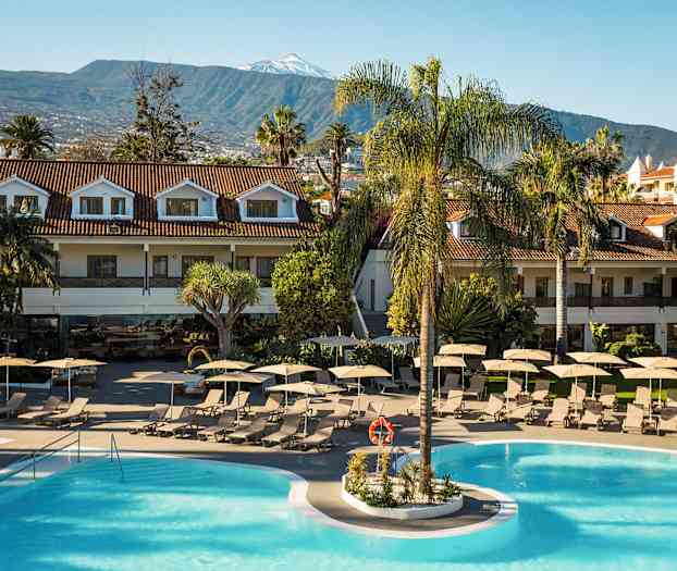 Lomakeskus uima-allasalueen kanssa, ympäröitynä aurinkotuoleilla ja -varjoilla. Vieressä rakennuksia punaisilla katoilla ja vuoria taustalla. (Tekoälyn luoma teksti)