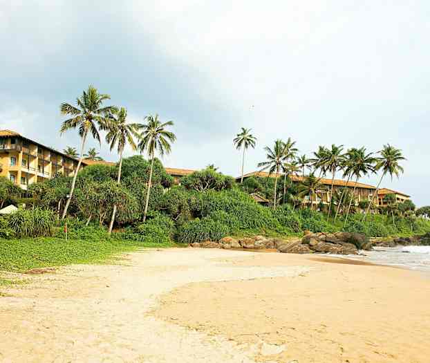 Hiekkaranta, taustalla vihreää pensaikkoa ja palmuja. Oikealla näkyy keltaseinäinen rakennus. (Tekoälyn luoma teksti)