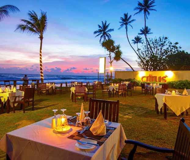 Ulkona sijaitseva ravintola valkoisine pöytäliinoineen ja puutuoleineen auringonlaskun aikaan merenrannalla. (Tekoälyn luoma teksti)