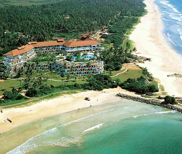 Ilmakuva hotellialueesta vehreyden ympäröimänä rannalla, jossa on pitkä hiekkaranta ja meri. (Tekoälyn luoma teksti)