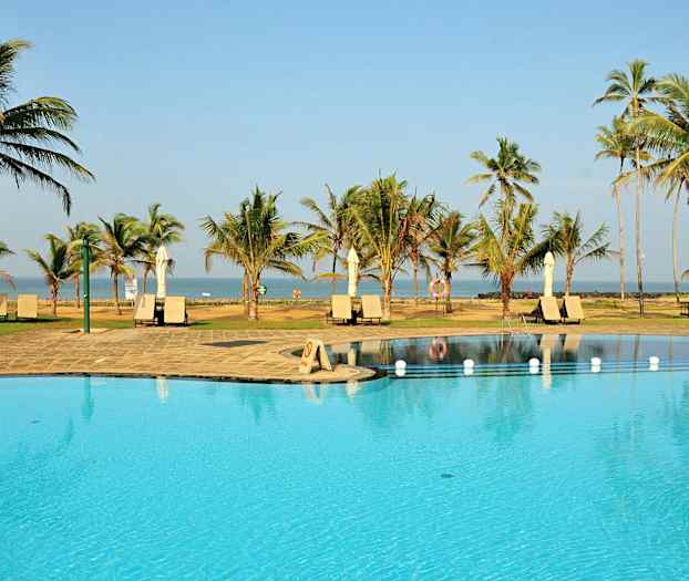 Kuva uima-altaasta, jossa on aurinkotuoleja ja varjoja trooppisessa paikassa, ympäröitynä palmuilla ja meri taustalla. (Tekoälyn luoma teksti)