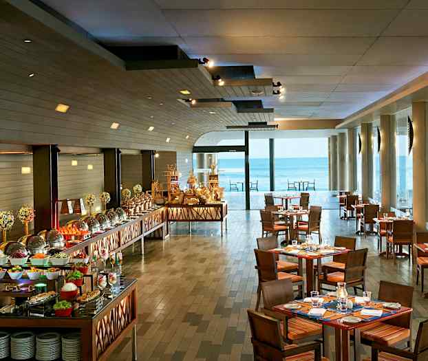 Kuva ravintolasta, jossa on katettuja pöytiä ja buffet, sisältäen erilaisia ruokia, suurien merelle aukeavien ikkunoiden äärellä. (Tekoälyn luoma teksti)