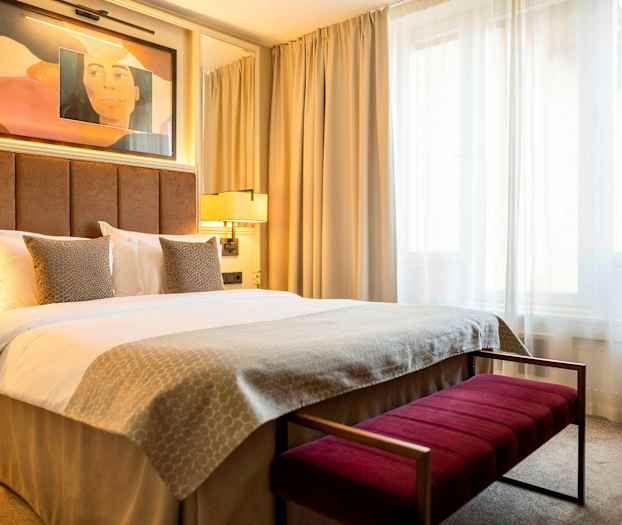 Hotellihuone, jossa on parivuode, beige vuodevaatteet ja kaksi tyynyä pehmustetun ruskean sängynpäädyn edessä. Taustalla näkyy valoja ja ikkuna vaaleilla verhoilla. (Tekoälyn luoma teksti)