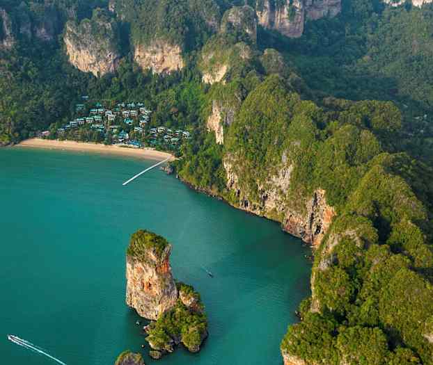 Ilmakuva trooppisesta rannikosta, jossa on ranta, kallioita ja vihreää kasvillisuutta sekä puita joukossa näkyy rakennuksia. (Tekoälyn luoma teksti)