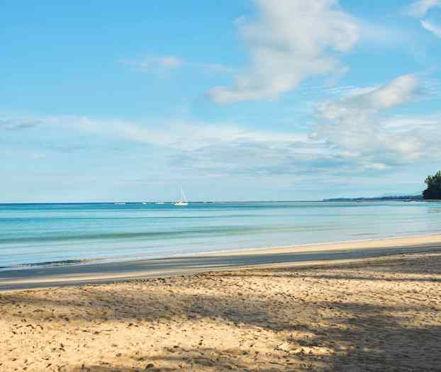 Hiekkaranta rauhallisella merinäkymällä, kaksi purjevenettä horisontissa ja puiden reunustamat rannat oikealla puolella. (Tekoälyn luoma teksti)