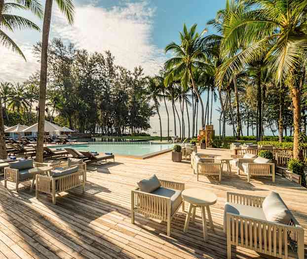 Puuterassi harmailla sohvaryhmillä ja sohvapöydillä, aurinkotuoleja uima-altaan ympärillä ja ympäristössä palmuja sekä puita. (Tekoälyn luoma teksti)