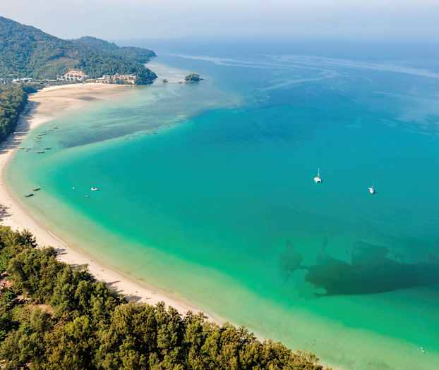 Pitkä valkoinen hiekkaranta turkoosinsinistä merta vasten, veneitä ja vehreitä ympäristöjä. (Tekoälyn luoma teksti)
