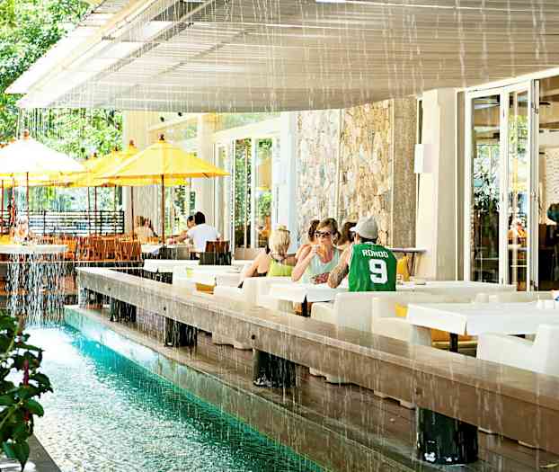 Ulkotila, jossa on pöytiä ja tuoleja, sekä vesikanava ja kelta-valkoiset aurinkovarjot. (Tekoälyn luoma teksti)