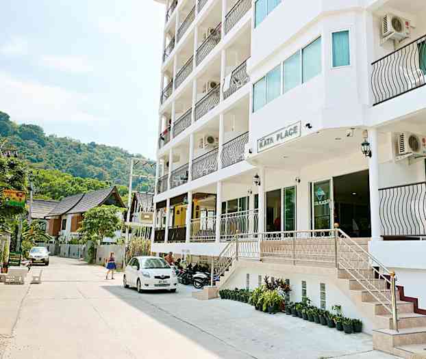 Kata Place -rakennuksen ulkopuolella on nykyaikainen rakennus parvekkeineen, pysäköityjä ajoneuvoja ja ohi kulkeva nainen. (Tekoälyn luoma teksti)