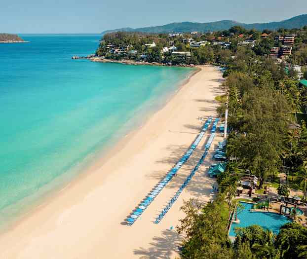 Ilmakuva hiekkarannasta, jossa on aurinkotuoleja ja varjoja pitkin merenrantaa sekä vihreitä alueita taustalla. (Tekoälyn luoma teksti)