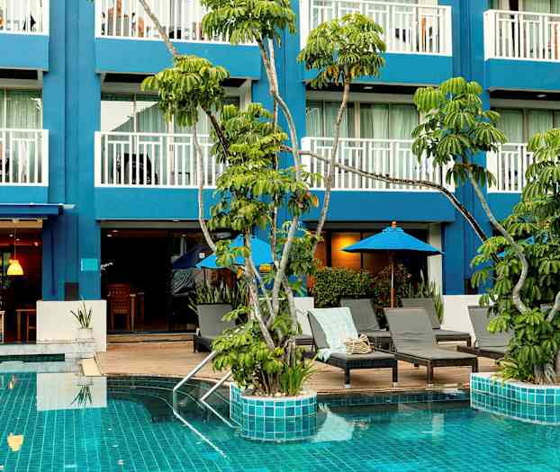 Kuvassa on uima-allas, jossa on turkoosi vesi, ympärillä aurinkotuoleja ja aurinkovarjoja sekä taustalla sininen rakennus parvekkeineen. (Tekoälyn luoma teksti)